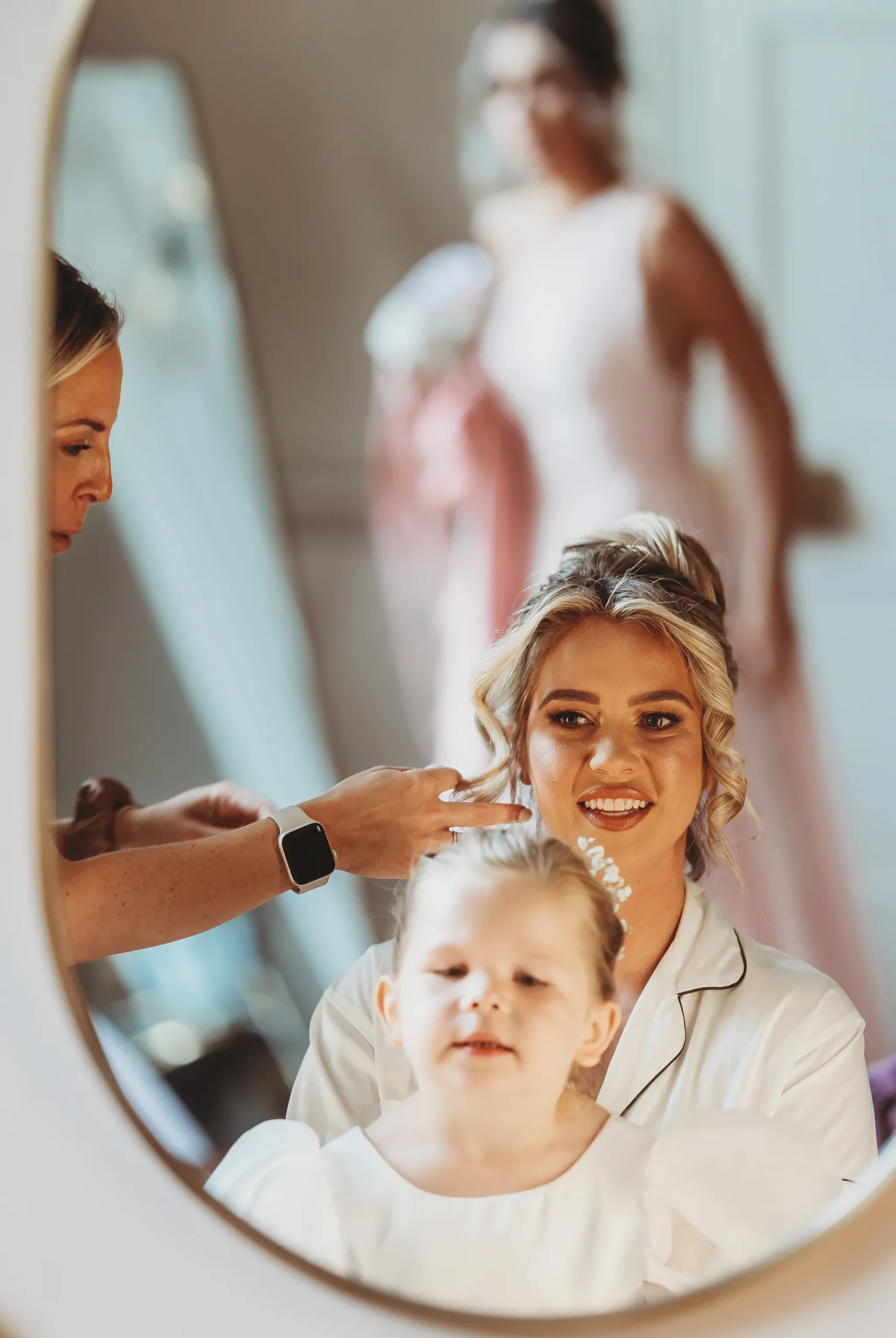 A bride having her hair finished, photographed into an arched mirror with a flower girl and bridesmaid, unposed wedding photography
