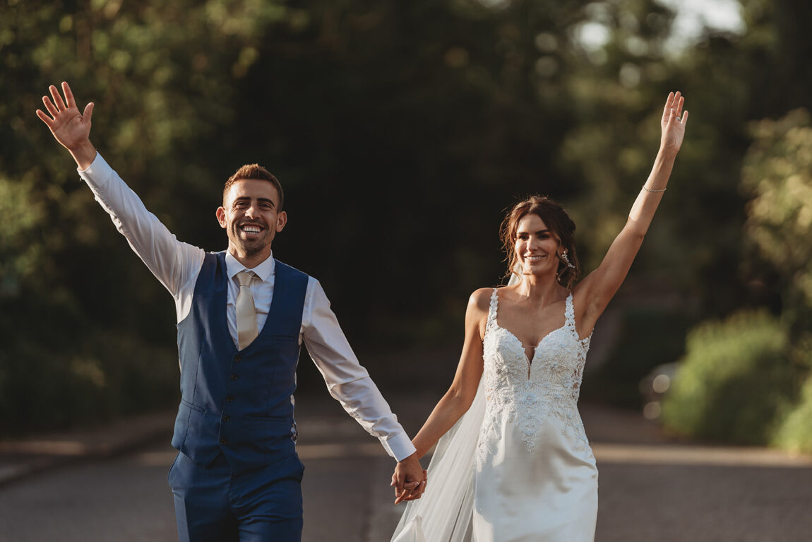 Bride and groom walking with arms in the air in evening light, Dedham