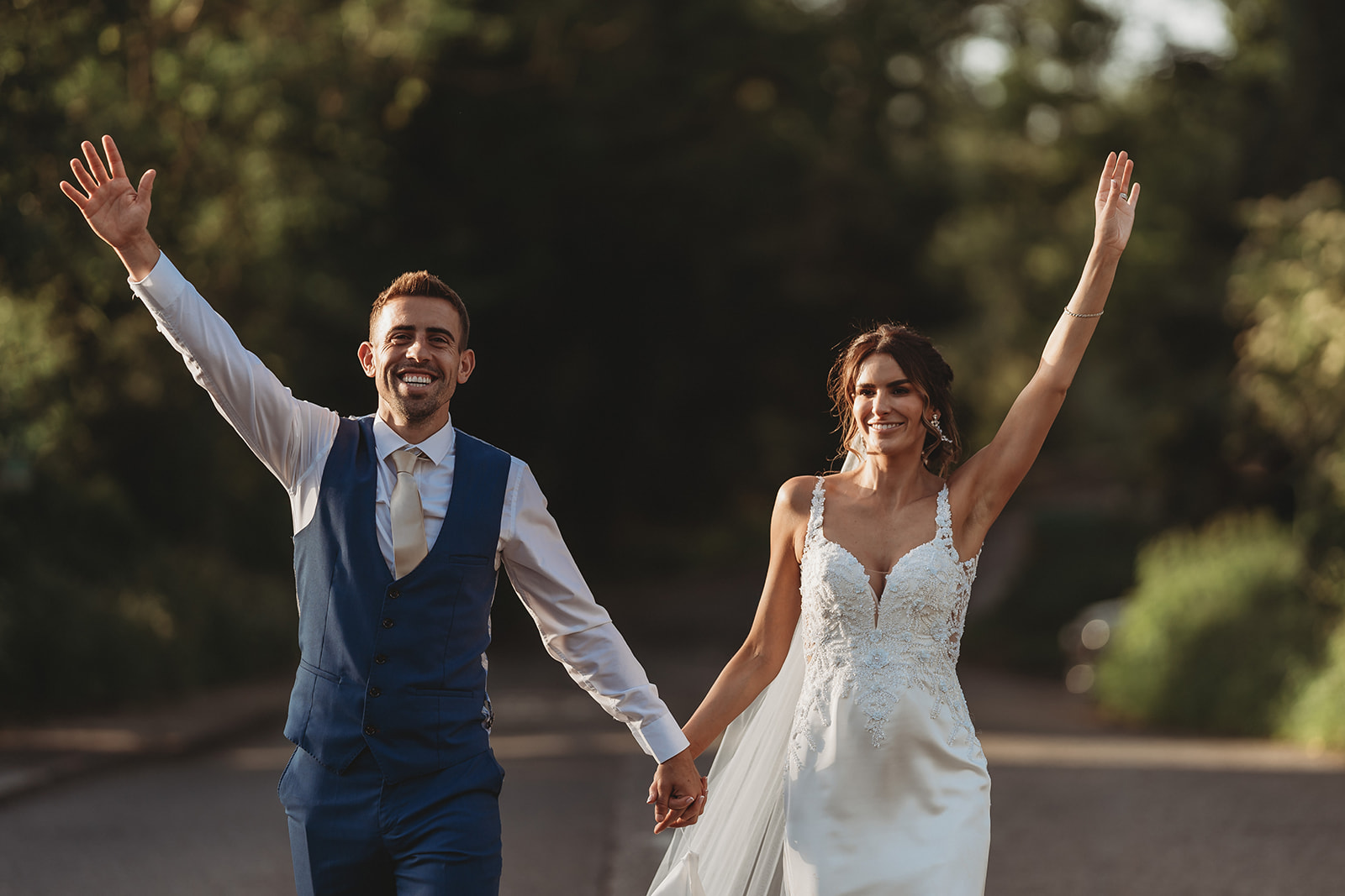 Bride and groom walking with arms in the air in evening light, Dedham