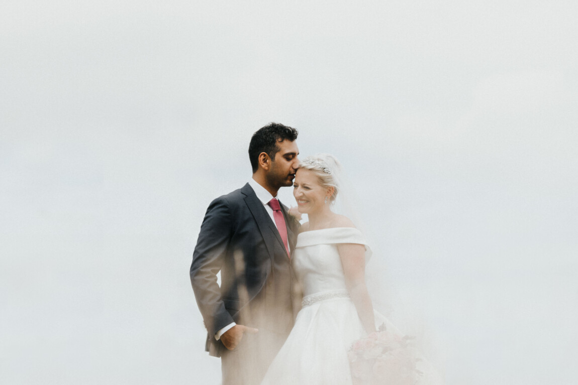 documentary wedding photography — groom kissing the bride’s forehead as her veil blows in the wind